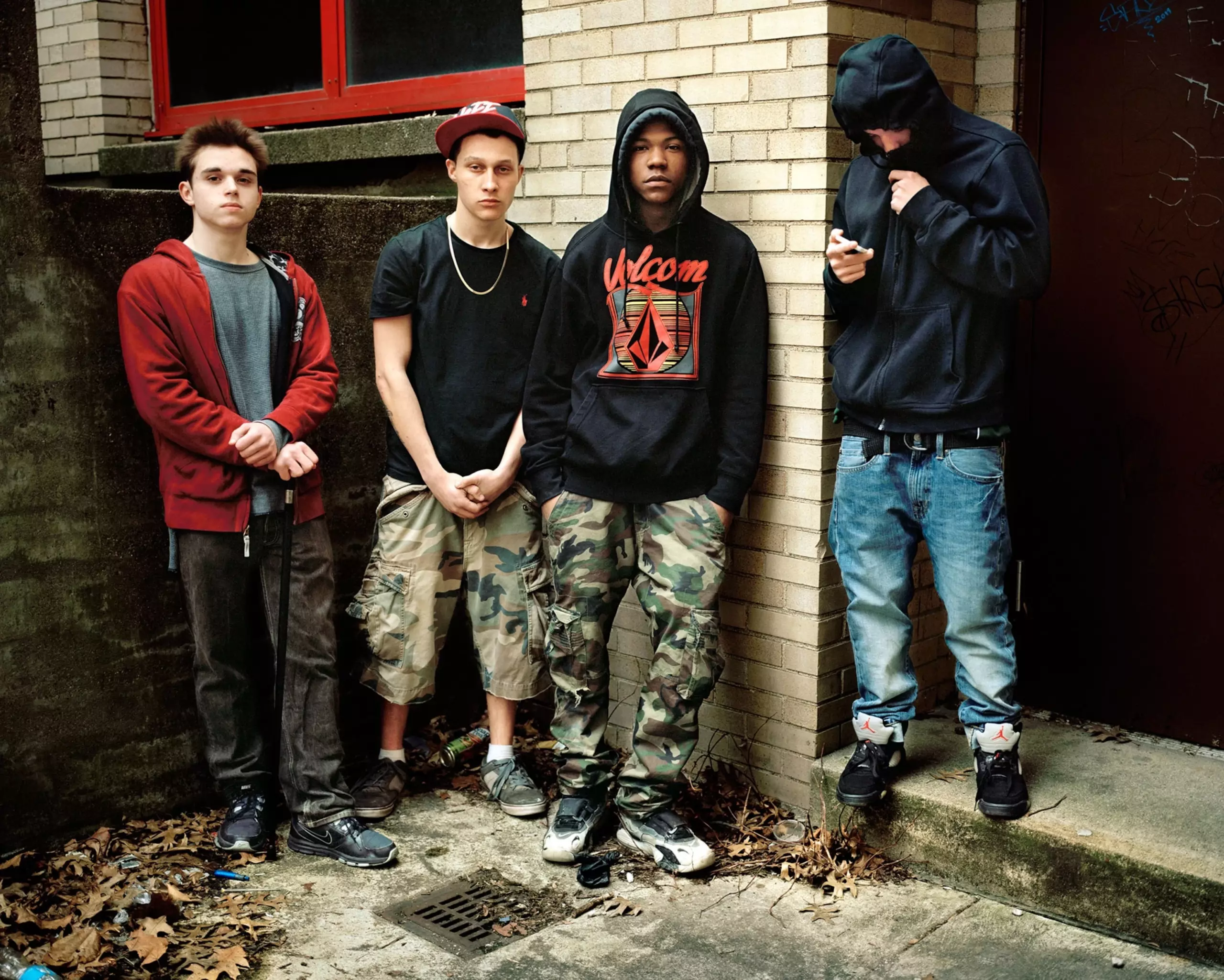 Four teenage boys dressed in hoodies, three of them standing and staring at the camera, one looking down at his phone